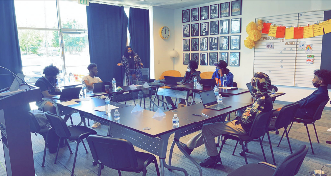 A group of students seated at tables with laptops while an instructor speaks at the front of the classroom.