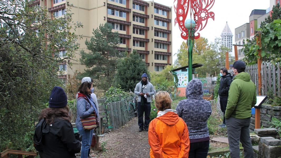 A small group gathers in an urban community garden, surrounded by trees, raised beds, and artwork, while a speaker addresses them. Tall residential and office buildings rise in the background, contrasting with the greenery and garden structures.