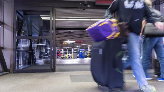 A blurred view of a person walking through an airport or transit terminal entrance while pulling a large wheeled suitcase and carrying another bag. Behind the moving person, there are glass doors, signs, and a row of parked shuttle vehicles visible in the background.