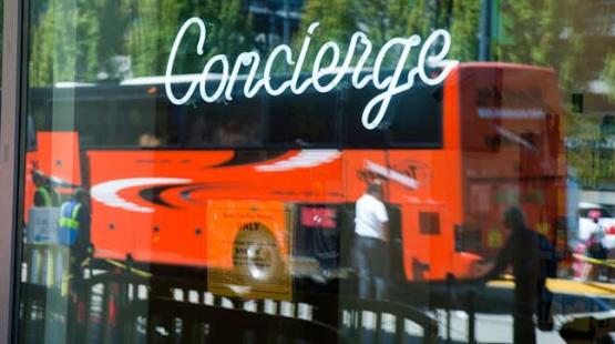 The word "Concierge" is written in white script on a reflective glass surface, through which a bright orange bus and several indistinct people standing nearby can be seen. The reflection shows outdoor trees and structures, creating a layered scene of signage and transit activity.