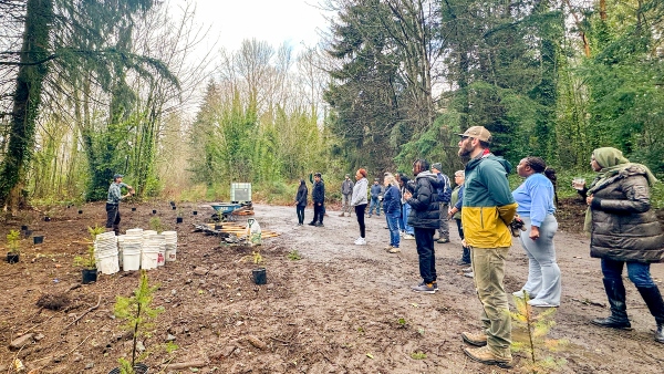 A group of people standing in a forest clearing listening to an instructor near buckets, tools, and small saplings prepared for planting.