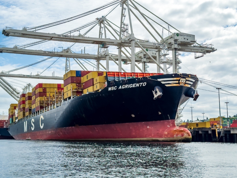 The container ship MSC Agrigento is docked at a port, loaded with stacked yellow and red cargo containers beneath large cranes.