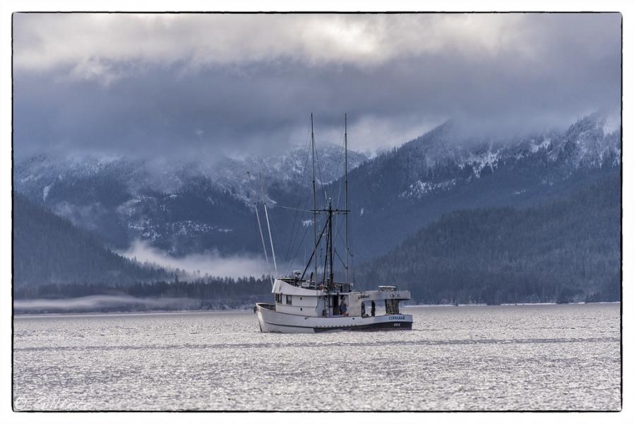 A salmon longliner sits on calm water with snow‑covered mountains and clouds in the background.