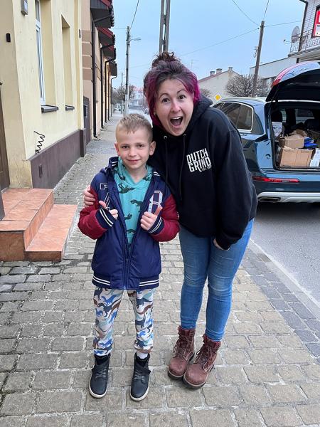 Two people standing on a sidewalk beside a building and an open vehicle with boxes inside. One person is standing slightly behind the other, and both are looking toward the camera.