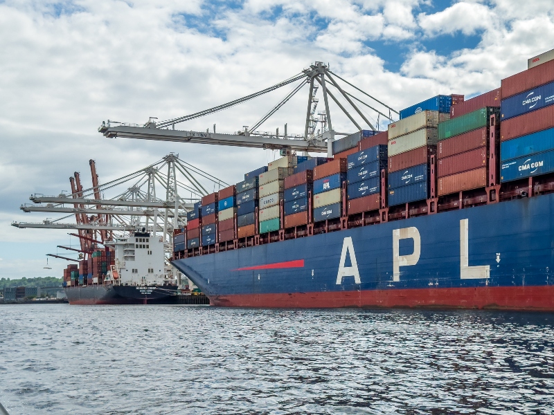 The container ship APL Charleston is docked at a port, loaded with stacked cargo containers beside large cranes.