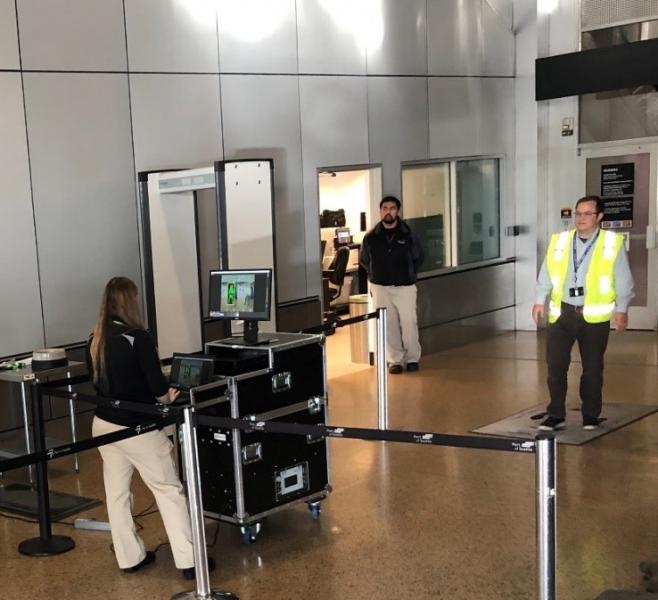 Security screening area with personnel operating a scanning device and a person walking through a monitoring zone.