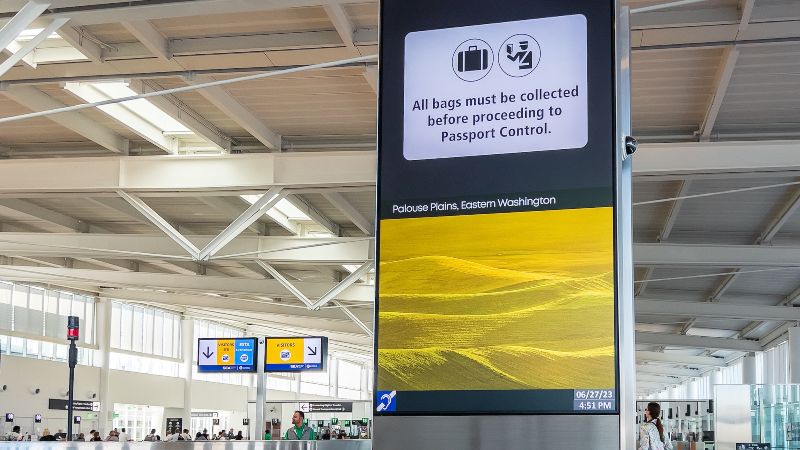 An airport terminal sign displays a message about collecting bags before passport control, with additional wayfinding signs visible in the background.