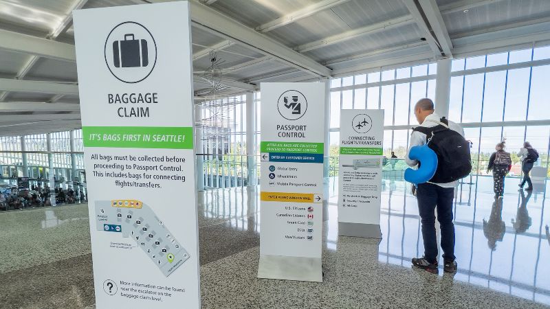 Signs for baggage claim, passport control, and connecting flights are displayed in an airport area, with a traveler standing nearby.