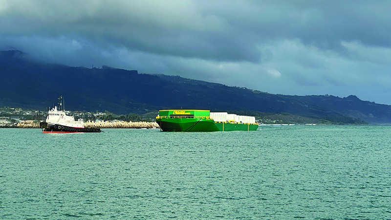 A tugboat guides a large green and yellow cargo barge carrying white‑wrapped freight near a coastline under cloudy skies.