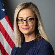 Marsiha Beganovic wearing glasses and a dark top, posed in front of a gray background with part of a U.S. flag visible.