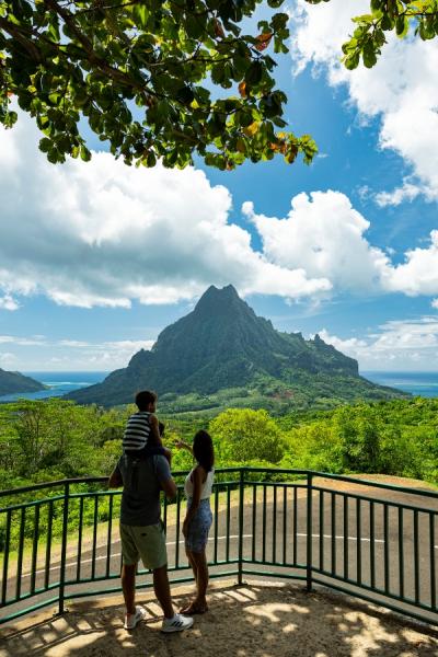 Three people stand at a scenic overlook facing a tall, lush green mountain with ocean visible in the distance.