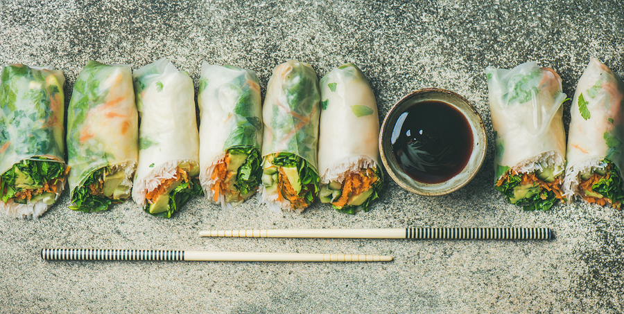 A row of fresh vegetable spring rolls arranged on a textured surface, with a small bowl of dark dipping sauce and a pair of wooden chopsticks placed in front.