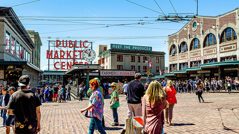 Crowds walk through an outdoor market area with the “Public Market Center” sign and clock visible.
