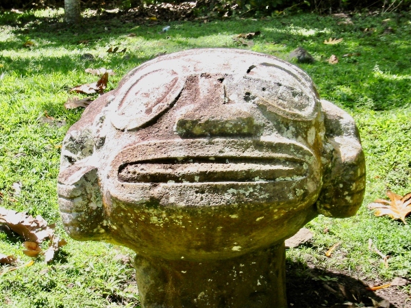 A weathered stone carving of a face with large circular eyes and a wide mouth, set outdoors on a grassy area.