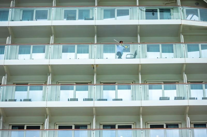 A worker cleans the glass railing of a cruise ship balcony deck, with rows of identical balcony cabins visible.