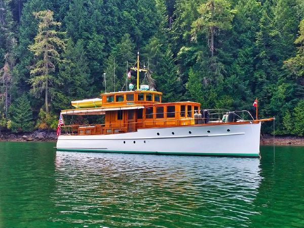 A classic white wooden motorboat named Bo‑Peep II floats on calm green water with a dense forest of tall evergreen trees in the background. The vessel features varnished wood cabin structures, a small yellow tender secured on the roof, and several flags on its mast. An American flag flies at the stern.