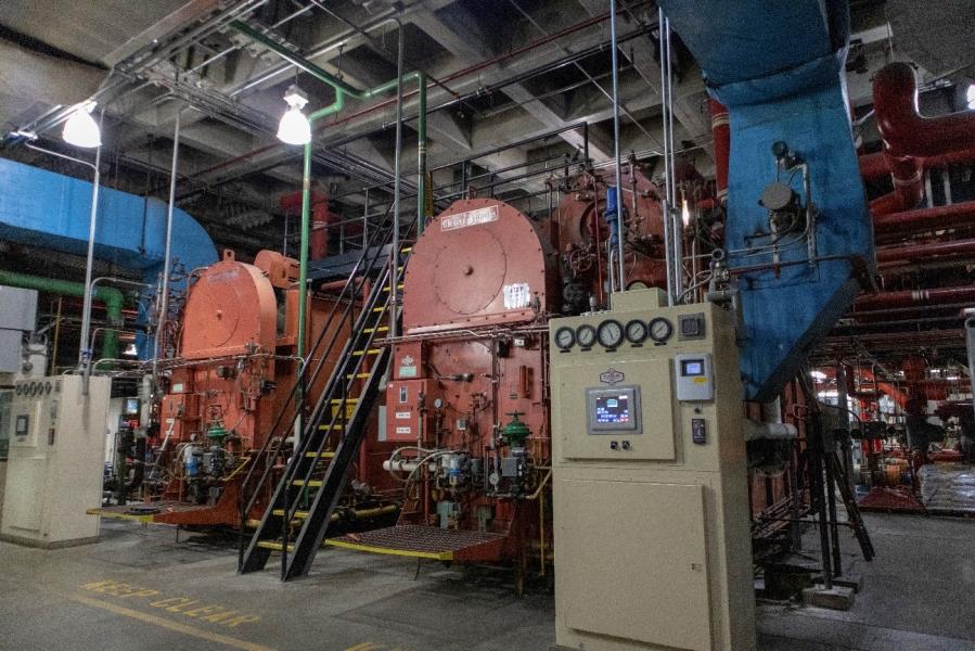 Large industrial boilers and mechanical equipment inside a utility plant, with control panels, pipes, and metal stairways.
