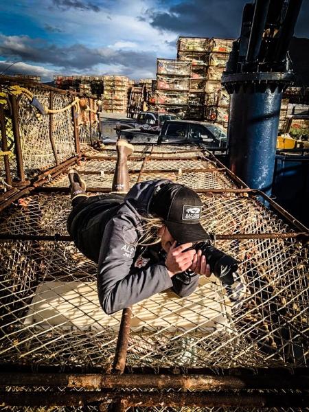 Bri Dwyer wearing outdoor gear lies on top of a large metal crate, using a camera, with stacks of equipment visible in the background.
