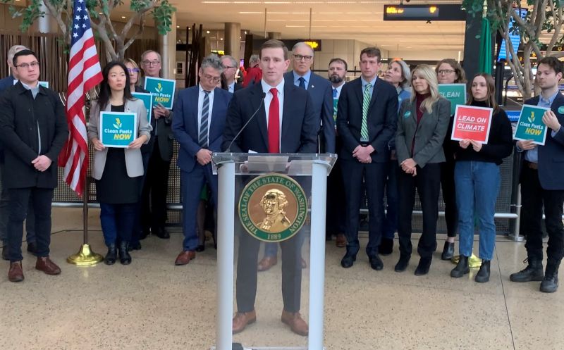 Port of Seattle Commissioner Ryan Calkins speaking at a podium in an airport terminal, with a group of people standing behind holding signs related to clean energy and climate.