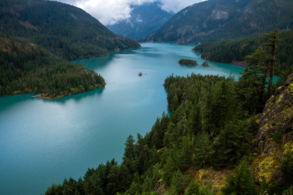 A long, blue reservoir winds between forested mountains under a cloudy sky.