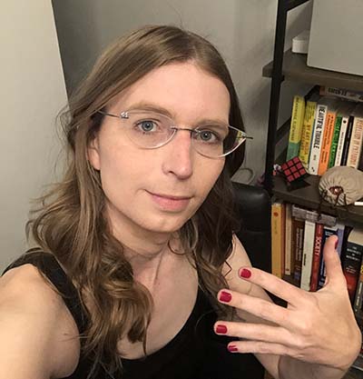 Chelsea Manning with long hair and glasses holds up a hand with painted fingernails while sitting indoors near a bookshelf.