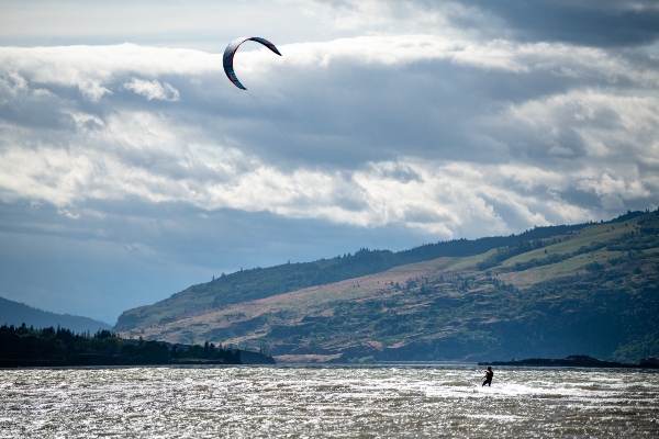 A person kiteboarding across choppy water beneath a cloudy sky with hills in the background.
