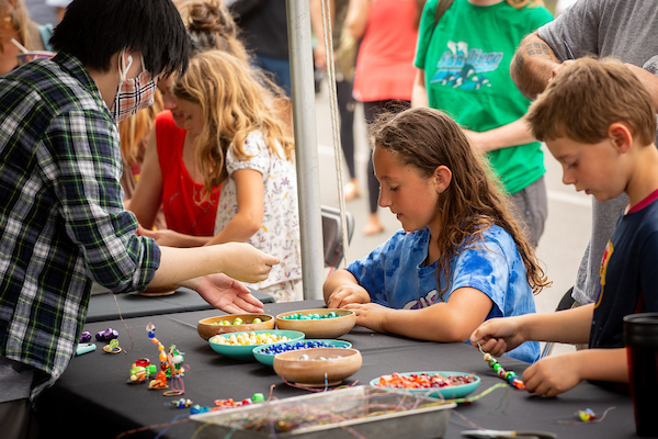 Children and an adult at a table creating crafts with colorful beads at an outdoor event.