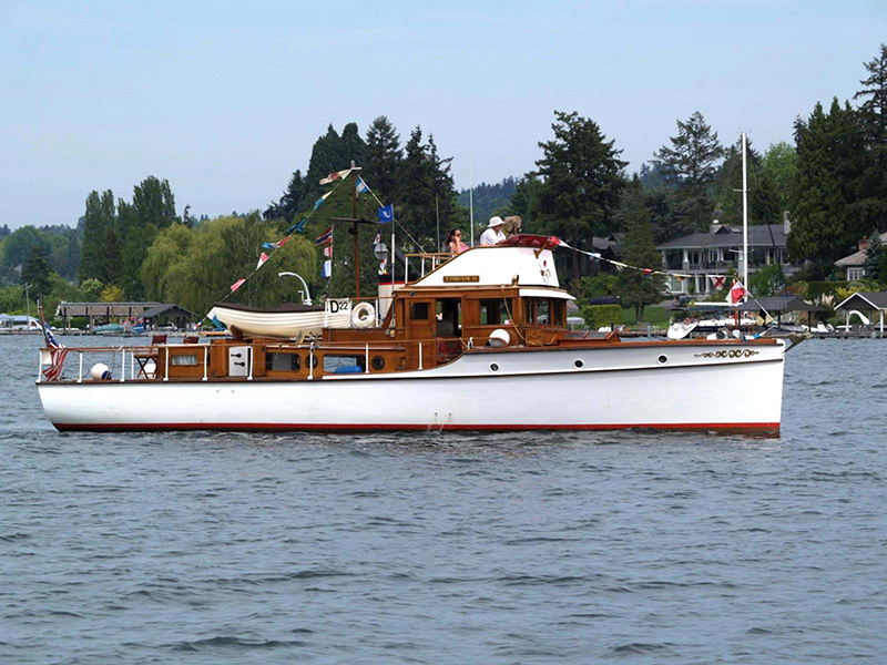 A classic white wooden motorboat named Corsair II moves across a calm lake with shoreline homes, trees, and moored boats in the background. The vessel features varnished wood trim, a small white tender secured on the roof, and colorful signal flags strung along the mast. Several people are visible on the upper deck.