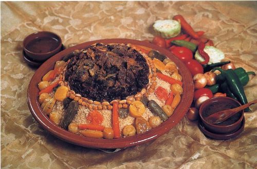 Large platter of couscous topped with stewed meat and surrounded by vegetables such as carrots, zucchini, and chickpeas, with bowls of sauce and fresh vegetables on the side.
