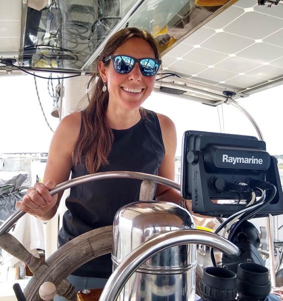 Esther Young at the helm of a boat holds the wheel near navigation equipment under a covered cockpit.