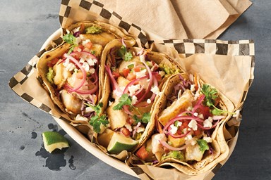 Three fish tacos filled with fried fish, vegetables, and garnishes, served in a paper-lined basket.