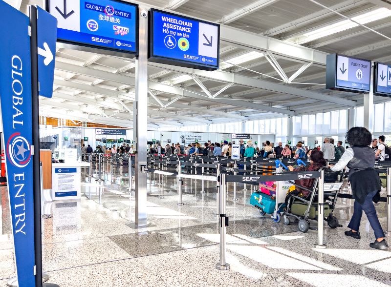 An airport security area with signage for Global Entry, wheelchair assistance, and flight crew, with travelers moving through the queue.