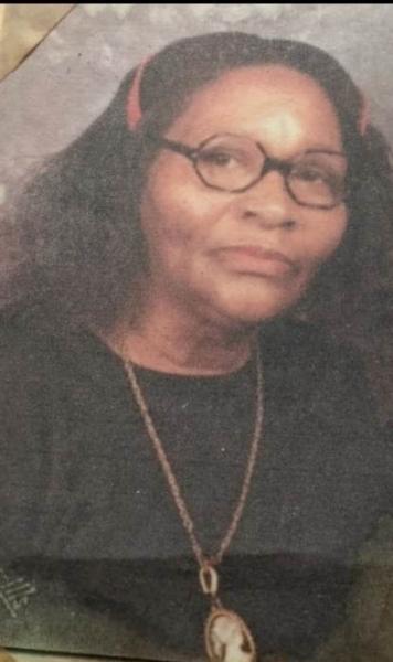 A portrait of Grandma Roena wearing glasses and a dark top with a pendant necklace, posed against a softly blurred background.