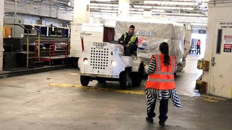 Airport service area where a small tow vehicle is pulling a large wrapped cargo container, with one worker walking nearby and another on the vehicle.