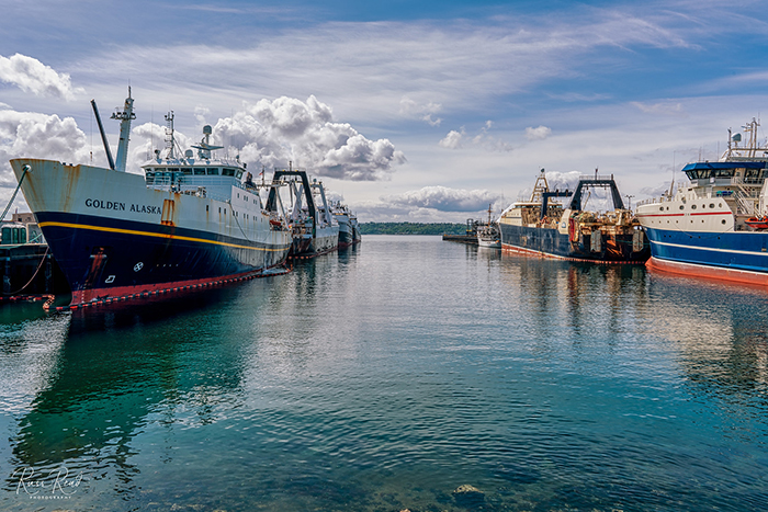 Fishing vessels lined up on both sides of a calm harbor under a partly cloudy sky.