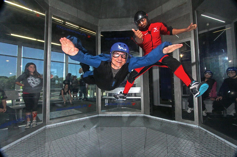 Two individuals wearing flight suits and helmets are floating in a vertical wind tunnel at an indoor skydiving facility, with several observers watching from behind the clear glass walls.