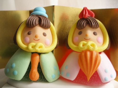 Two colorful, doll-like figurines with round faces, bright clothing, and decorative hoods, placed side by side against a gold background.