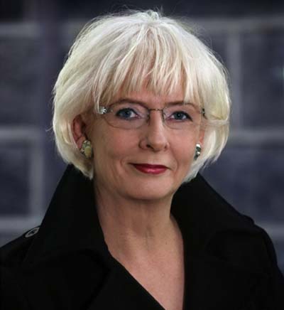 Jóhanna Sigurðardóttir with short, light-colored hair and glasses poses for a portrait while wearing a dark coat.