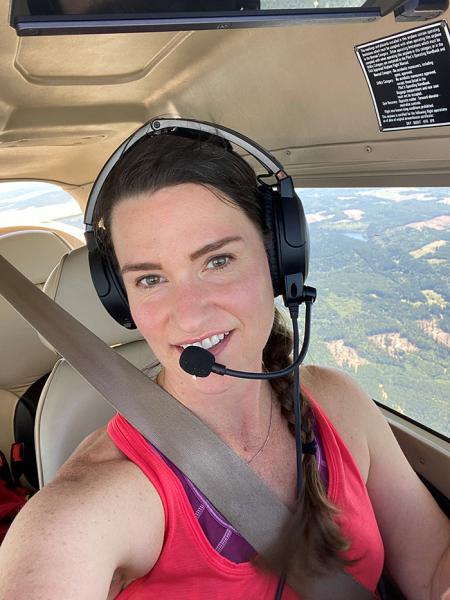 Katie Zera wearing a headset and seatbelt sits inside a small aircraft with a landscape visible through the window.