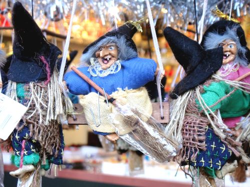 Three colorful hanging witch dolls with pointed hats, scruffy hair, and brooms, decorated with fabric clothing and displayed in a festive setting.