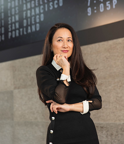 Luly Yang standing indoors in front of a flight information board, posed with one hand near the chin and wearing a long‑sleeved outfit with contrasting cuffs.