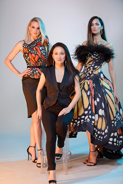 Luly Yang posing in a studio, with two wearing dresses featuring butterfly wing patterns and one seated in the center wearing a dark outfit.
