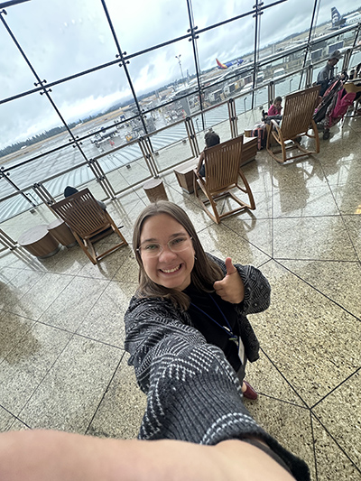 Alexa taking a selfie in an airport lounge area with large windows overlooking parked airplanes and seating.