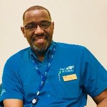 Amadou Ly wearing glasses and a blue collared shirt with an ID lanyard, seated in front of a plain light-colored wall.