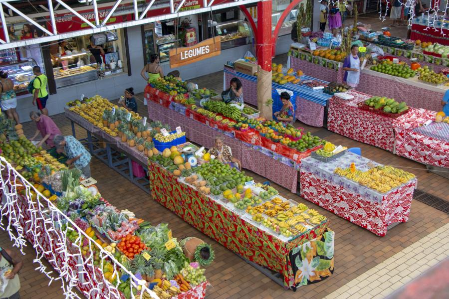 A marketplace with multiple tables covered in colorful cloths displaying fruits and vegetables, with shoppers and vendors moving between the stalls.