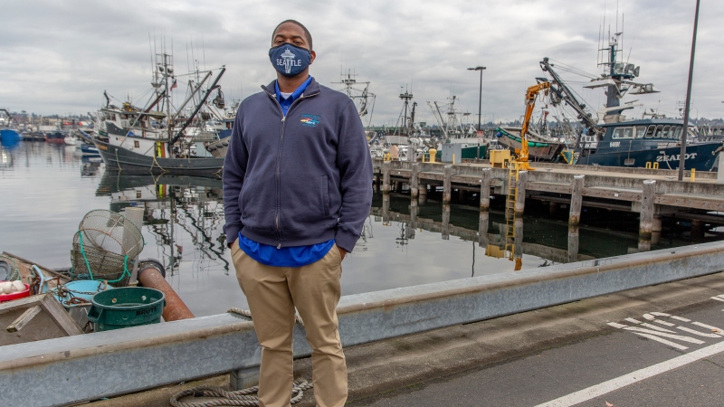 Port of Seattle employee wearing a face mask stands near a harbor with multiple fishing vessels docked in the background.