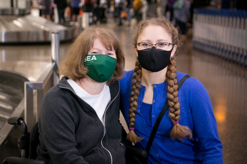 Two people wearing face masks pose together in an airport baggage claim area.