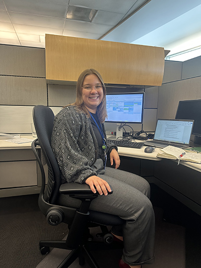 Alexa Meyer seated at an office workstation with multiple computer monitors and papers on the desk.