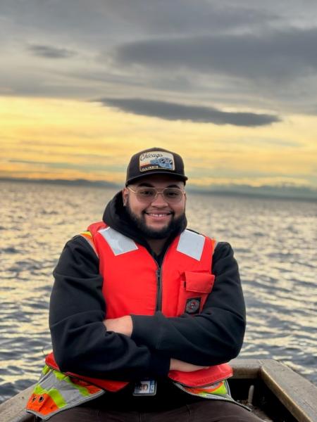 Michael Whiteside wearing a bright orange safety vest seated in a small boat on the water at sunset.