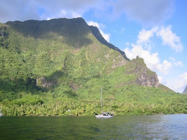 A sailboat floats on calm water below steep, lush green mountains under a partly cloudy sky.